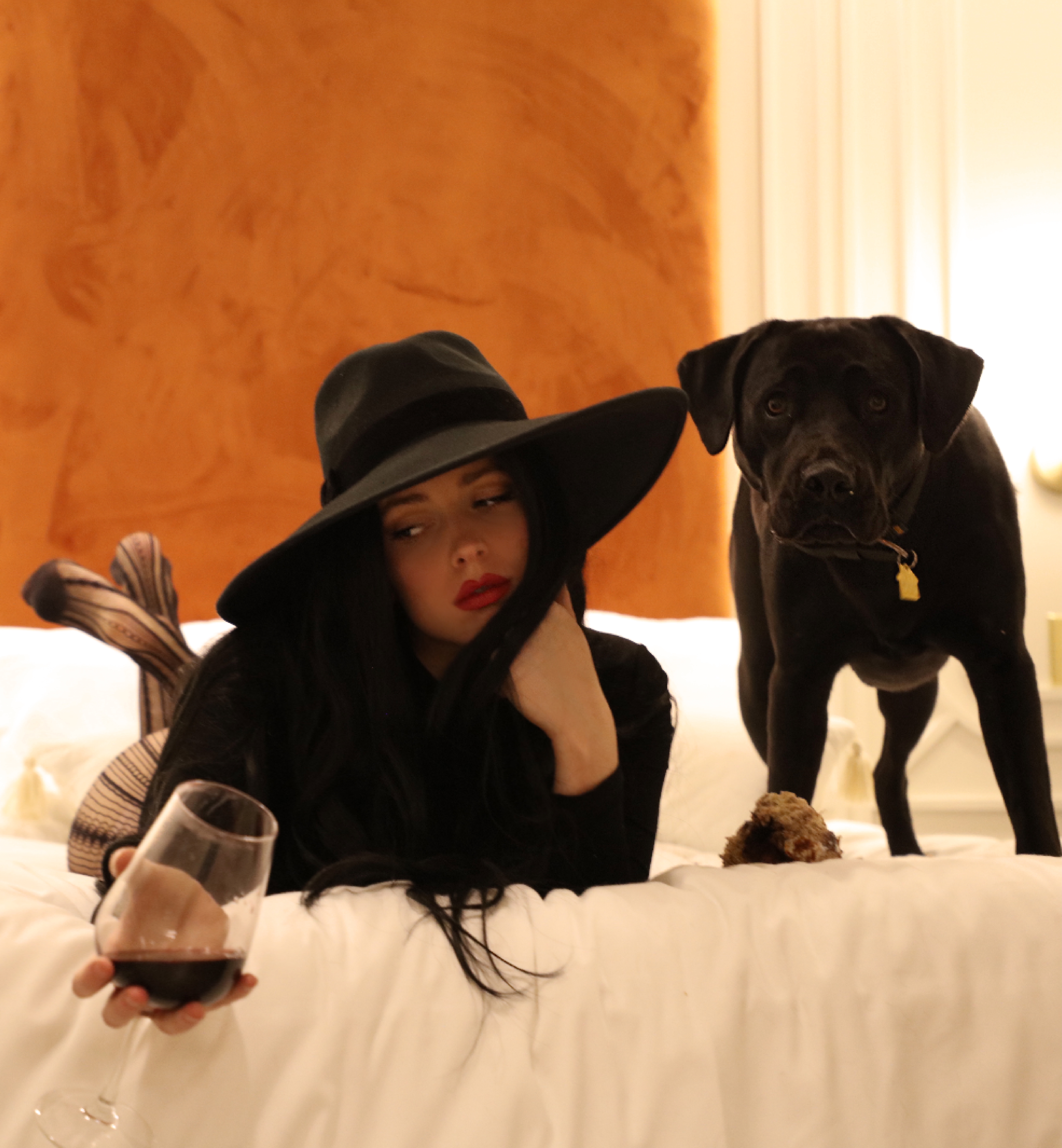 Merci Raines wearing a wide-brim black hat holding a wine glass and laying next to a black dog