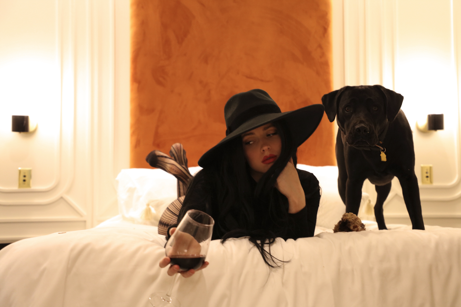 Merci Raines wearing a wide-brim black hat holding a wine glass laying next to a black dog